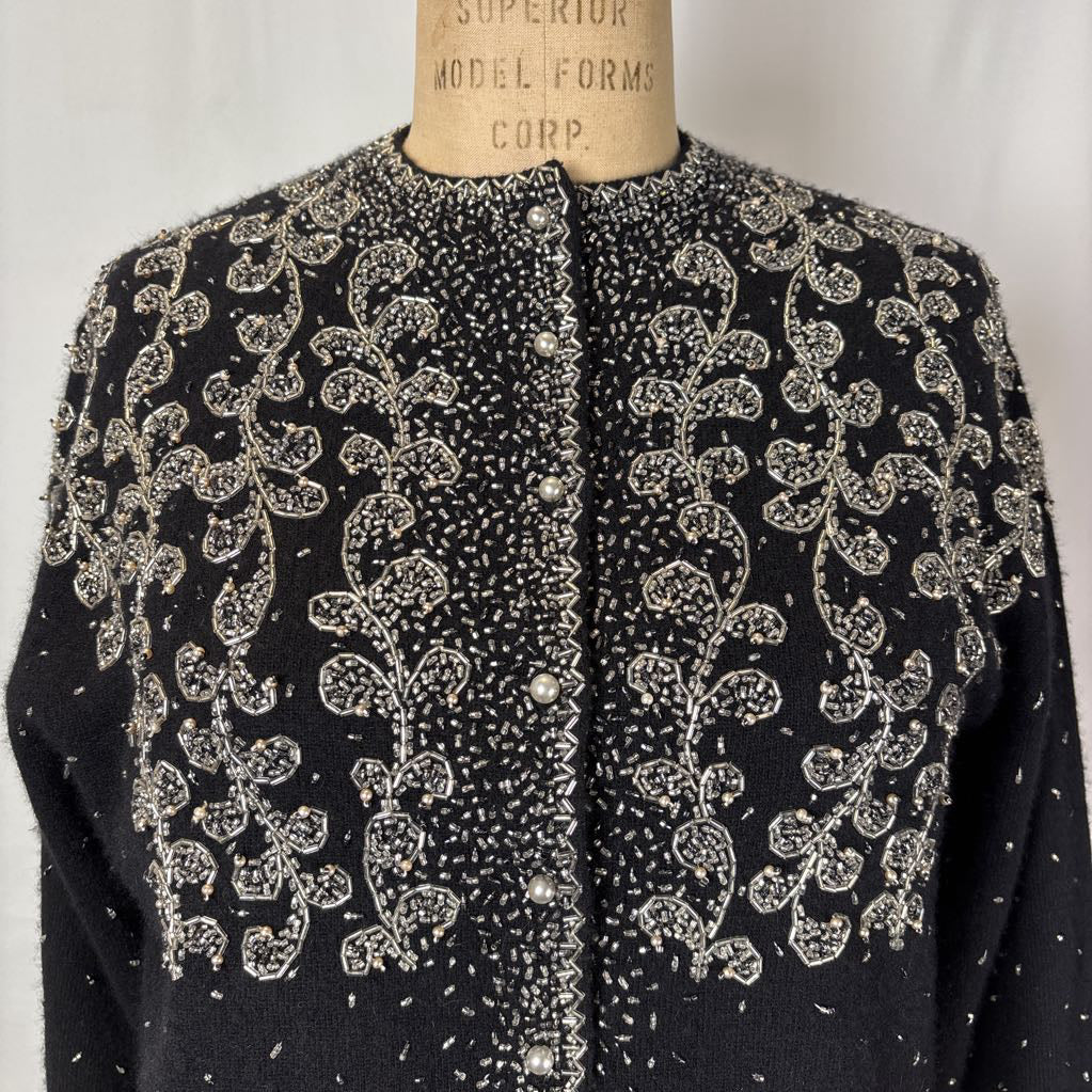 A black button-up sweater with intricate silver bead embroidery on an antique mannequin against a white wall. 