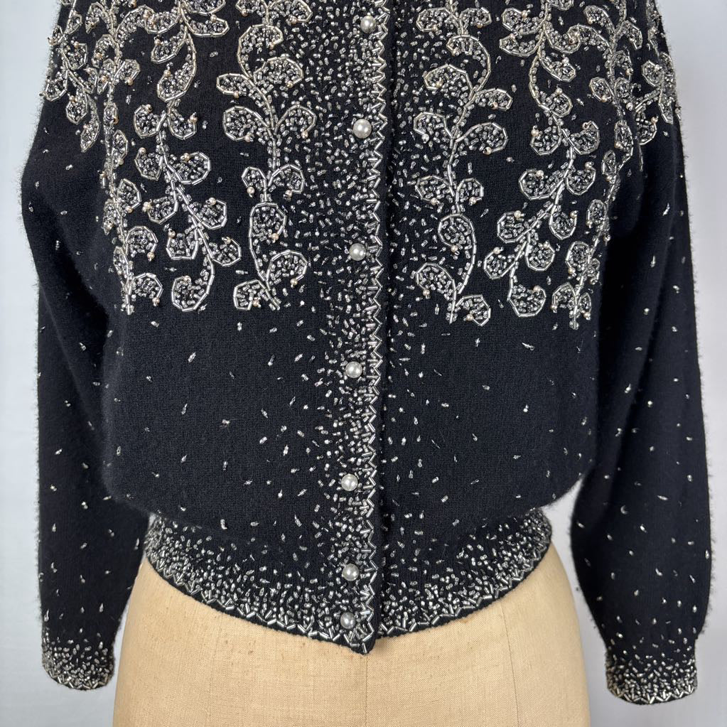 A black button-up sweater with intricate silver bead embroidery on an antique mannequin against a white wall. 