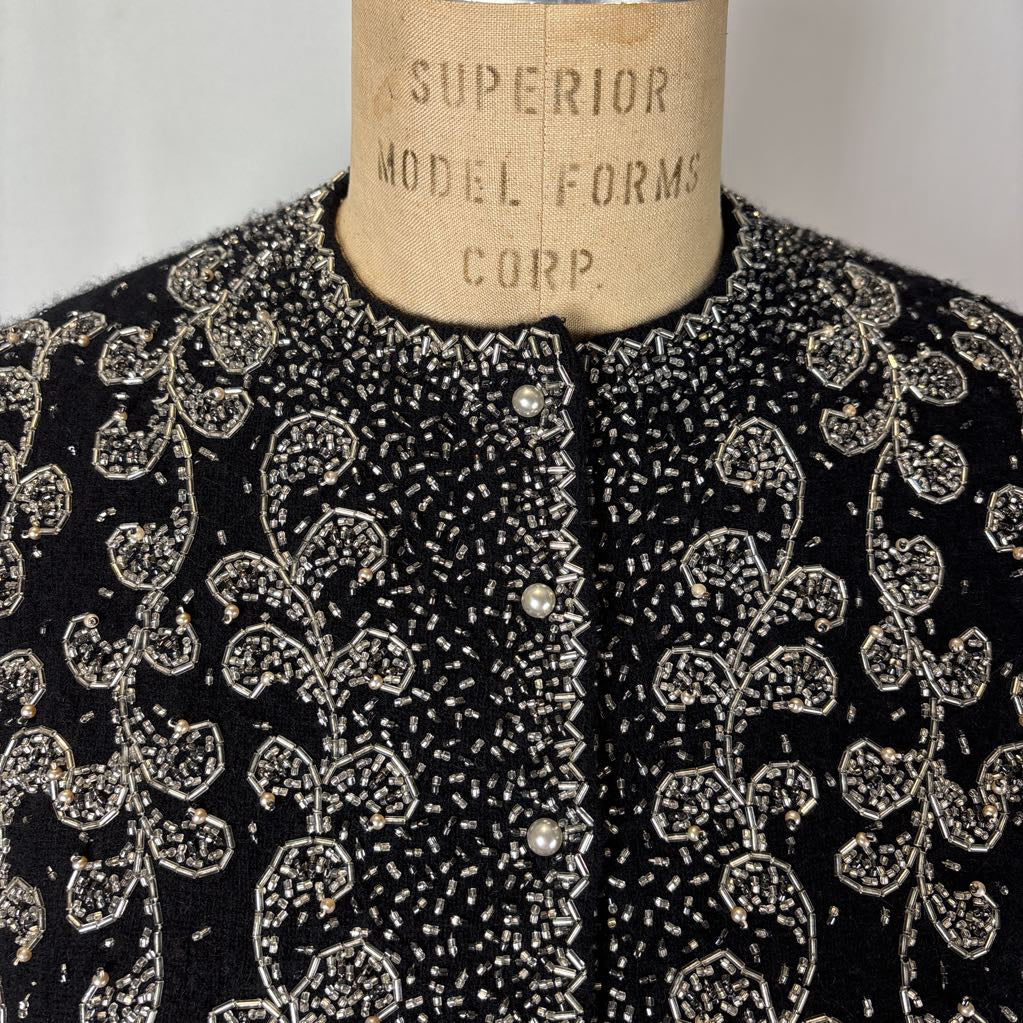 A black button-up sweater with intricate silver bead embroidery on an antique mannequin against a white wall. 