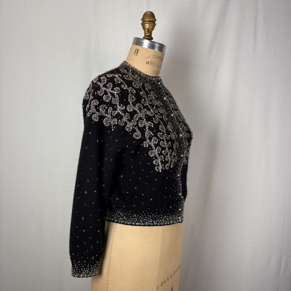 A black button-up sweater with intricate silver bead embroidery on an antique mannequin against a white wall. 