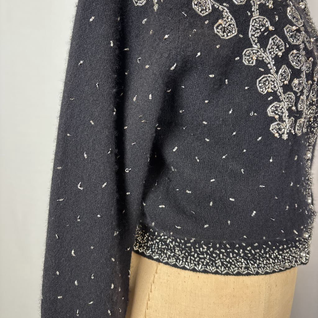 A black button-up sweater with intricate silver bead embroidery on an antique mannequin against a white wall. 