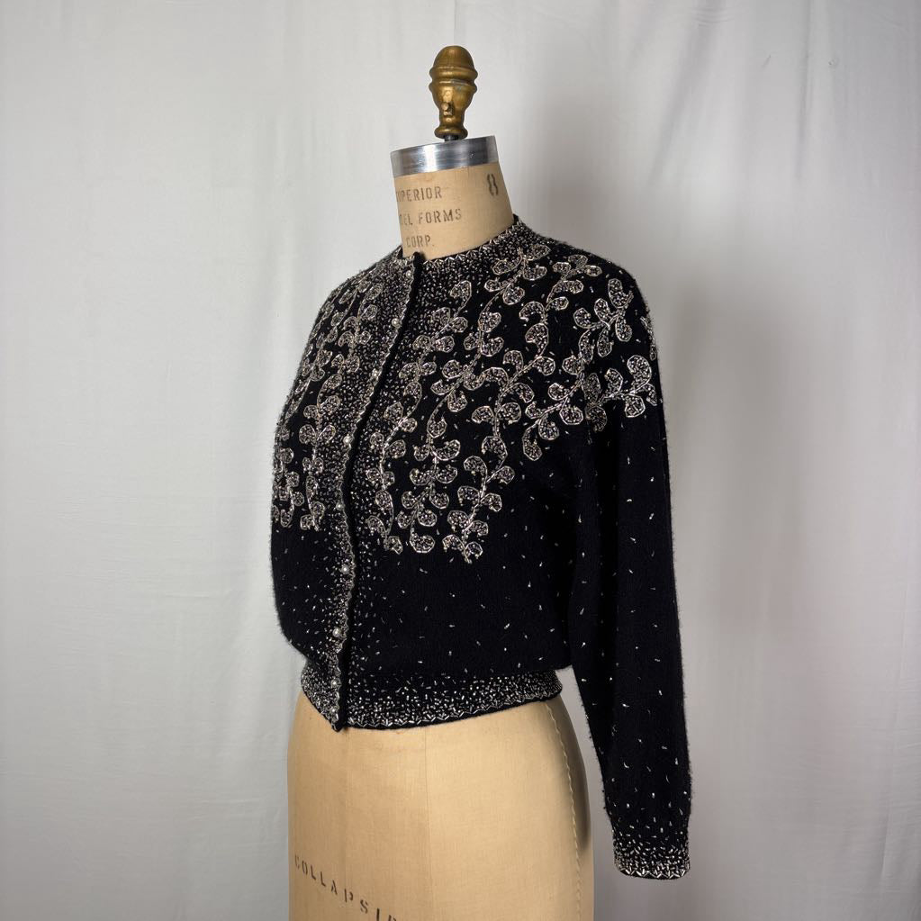 A black button-up sweater with intricate silver bead embroidery on an antique mannequin against a white wall. 