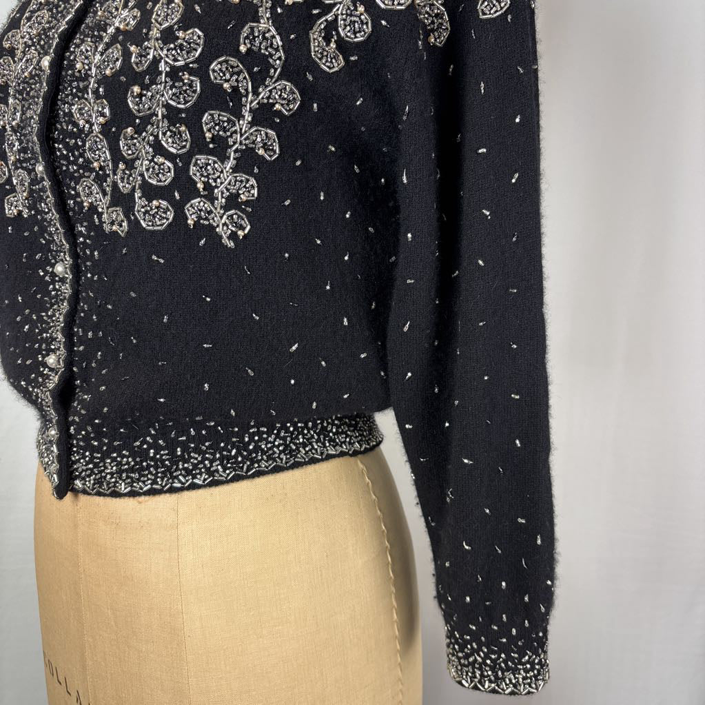 A black button-up sweater with intricate silver bead embroidery on an antique mannequin against a white wall. 