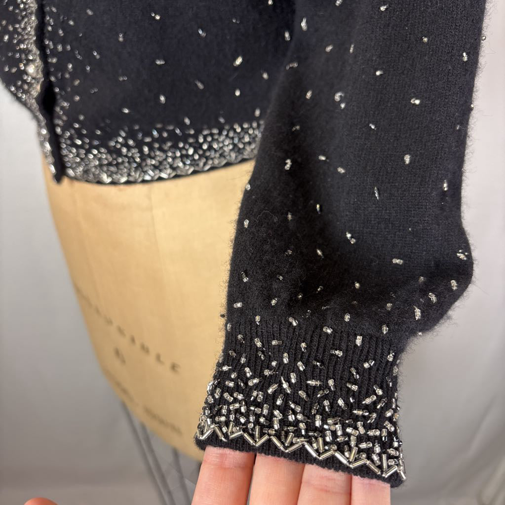 A black button-up sweater with intricate silver bead embroidery on an antique mannequin against a white wall. 