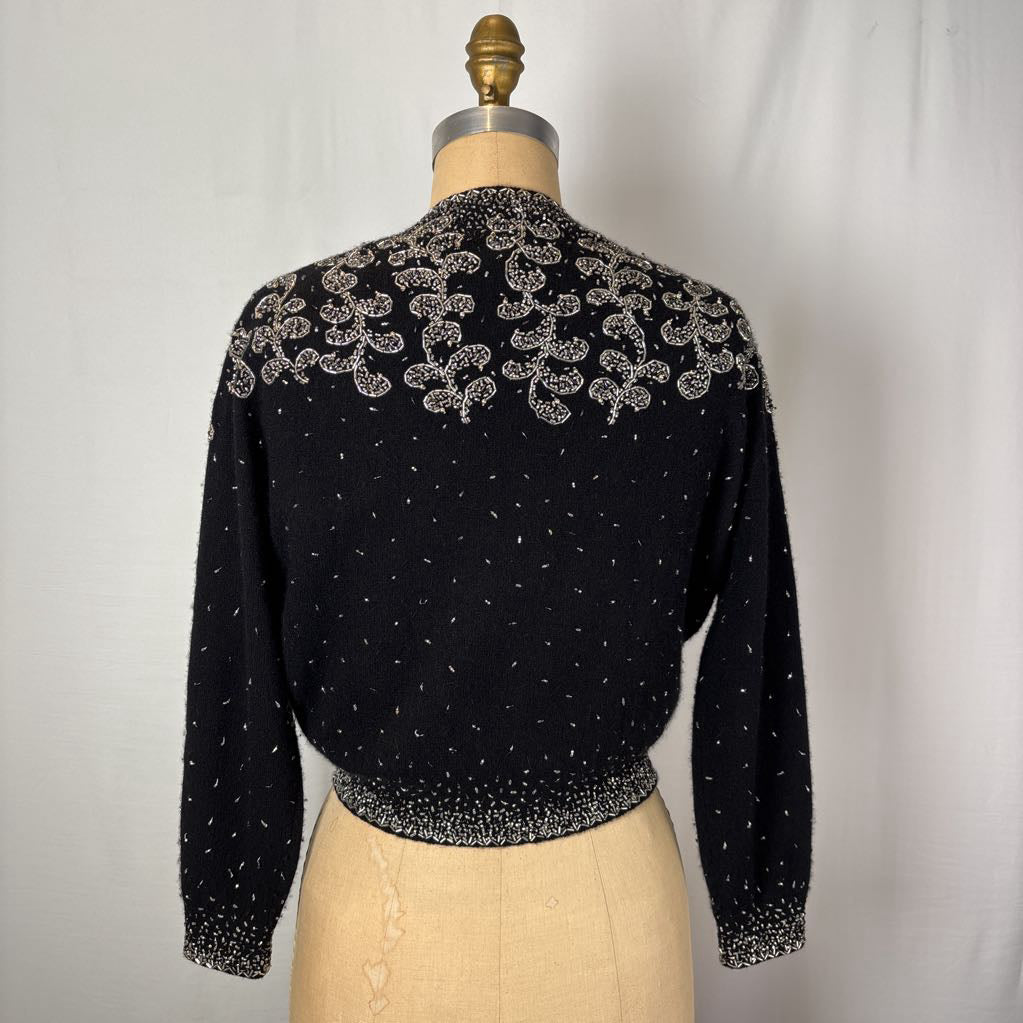 A black button-up sweater with intricate silver bead embroidery on an antique mannequin against a white wall. 