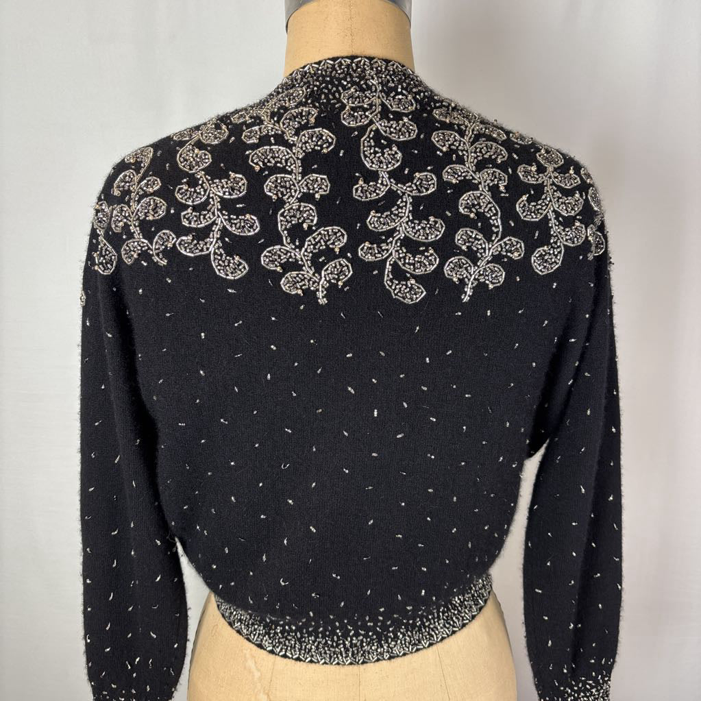 A black button-up sweater with intricate silver bead embroidery on an antique mannequin against a white wall. 