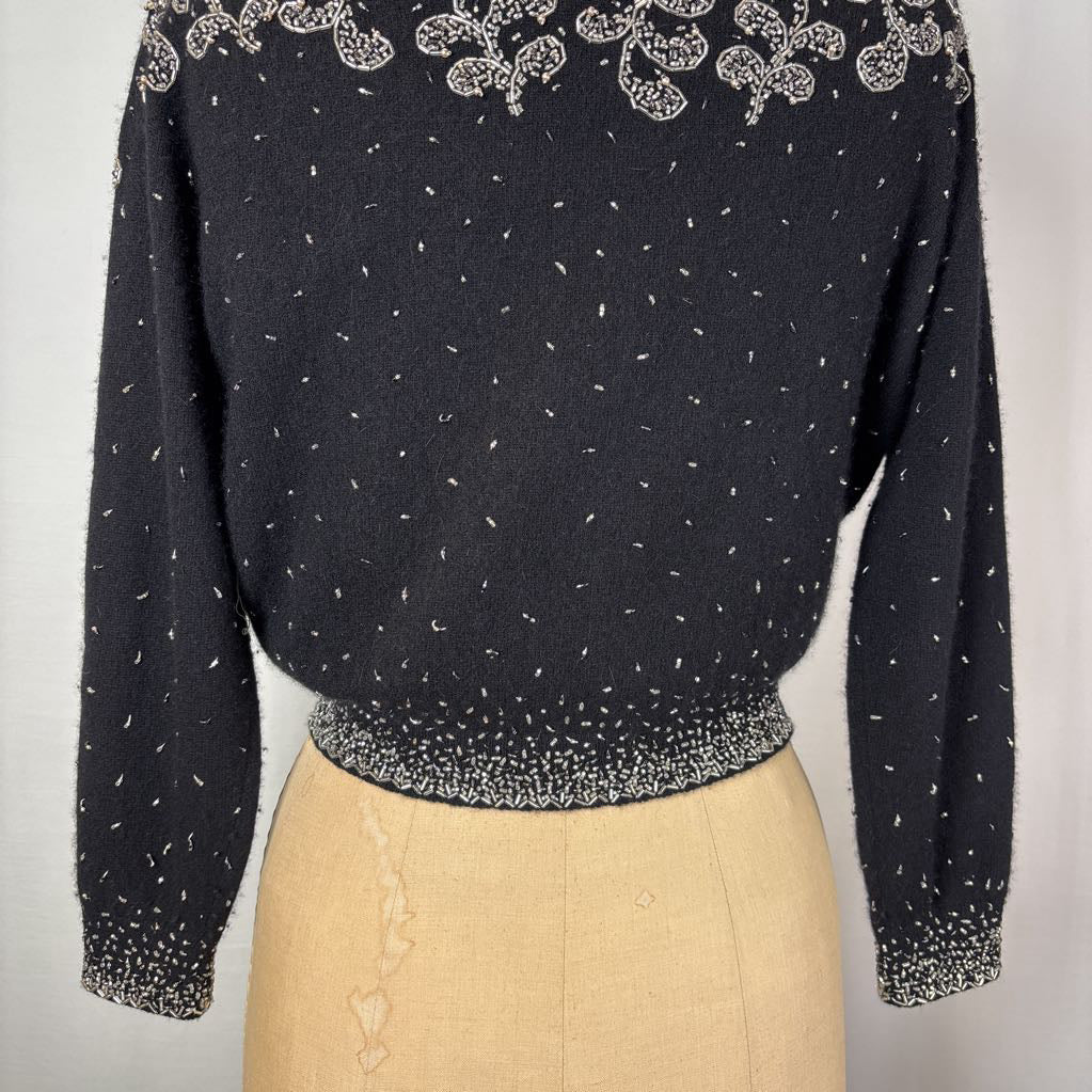 A black button-up sweater with intricate silver bead embroidery on an antique mannequin against a white wall. 