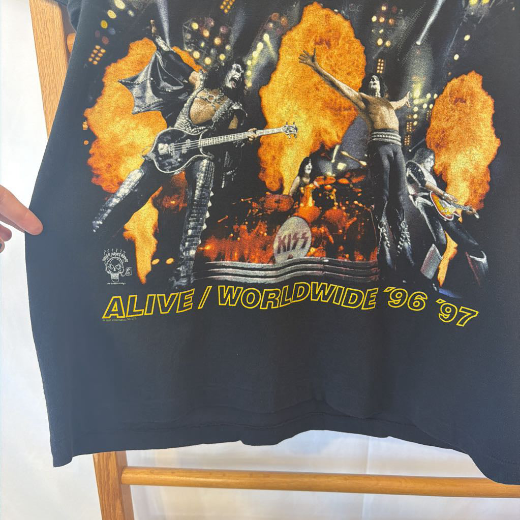 A black vintage Kiss 'Alive / Worldwide '96 '97 graphic T-shirt hanging on a wooden hanger. The shirt features a large front print of the band members performing with fiery orange and yellow flames, with the yellow text 'ALIVE / WORLDWIDE '96 '97' printed below.