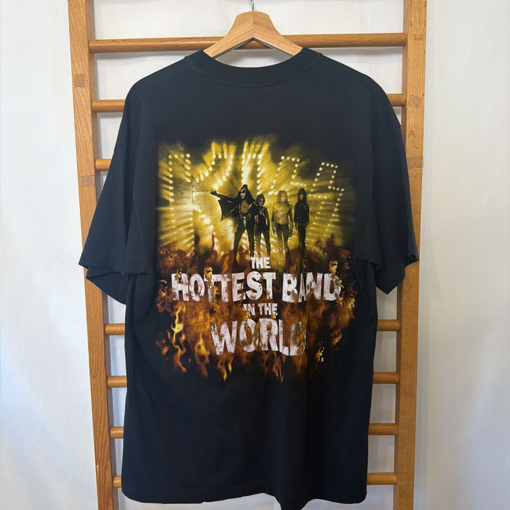 A black graphic t-shirt hanging on a wooden hanger against a white wall with a wooden ladder behind it. The shirt features a large yellow and orange concert-style graphic with the text 'THE HOTTEST BIRD IN THE WORLD'.