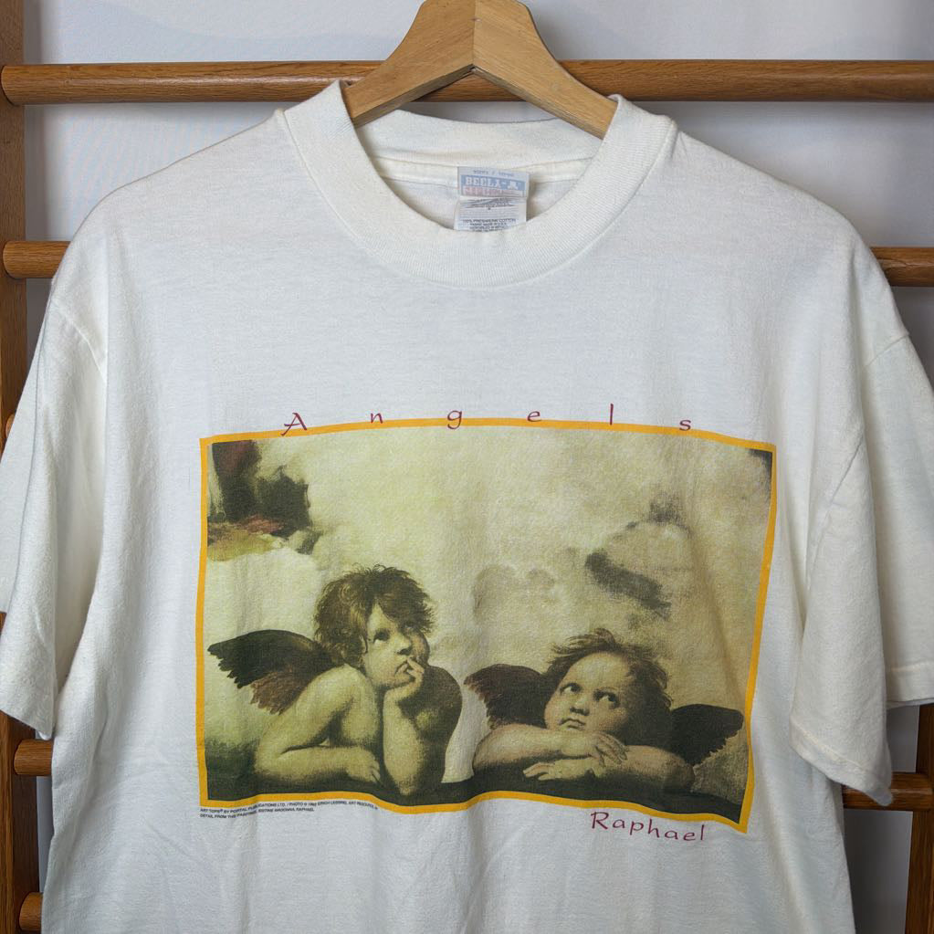 White t-shirt with angel graphic and 'Raphael' text on a wooden rack.