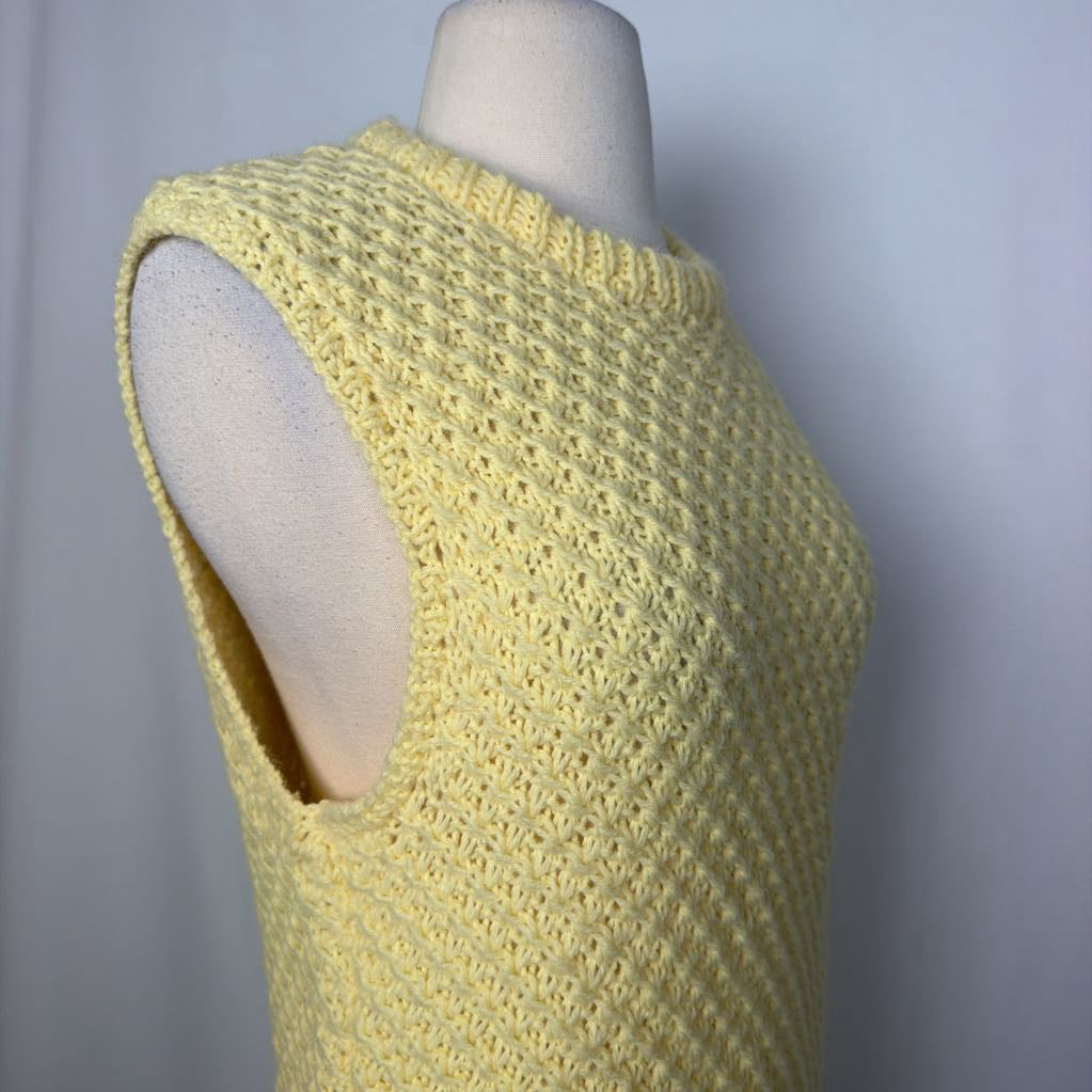 A pale yellow sleeveless knit top with a textured waffle pattern and a side cutout, displayed on a cream mannequin torso against a light gray background.