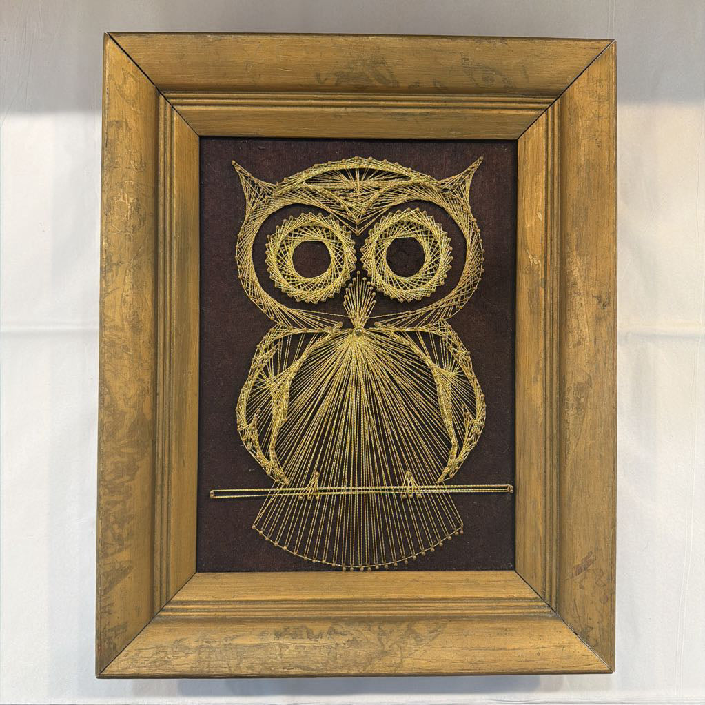 framed bronze owl  string art against a fabric brown background
against a white wall