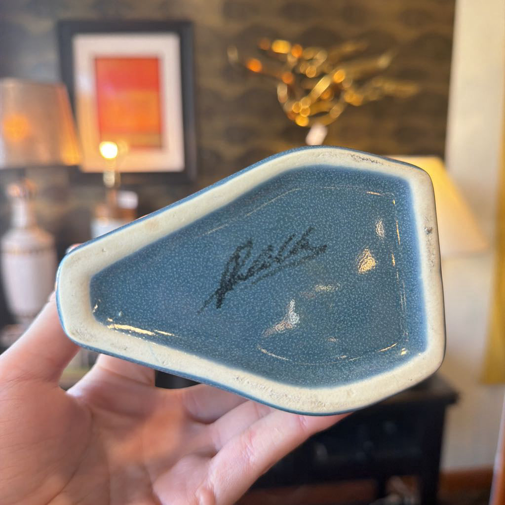 Hand holding a blue ceramic dish with a signature in a blurred indoor setting