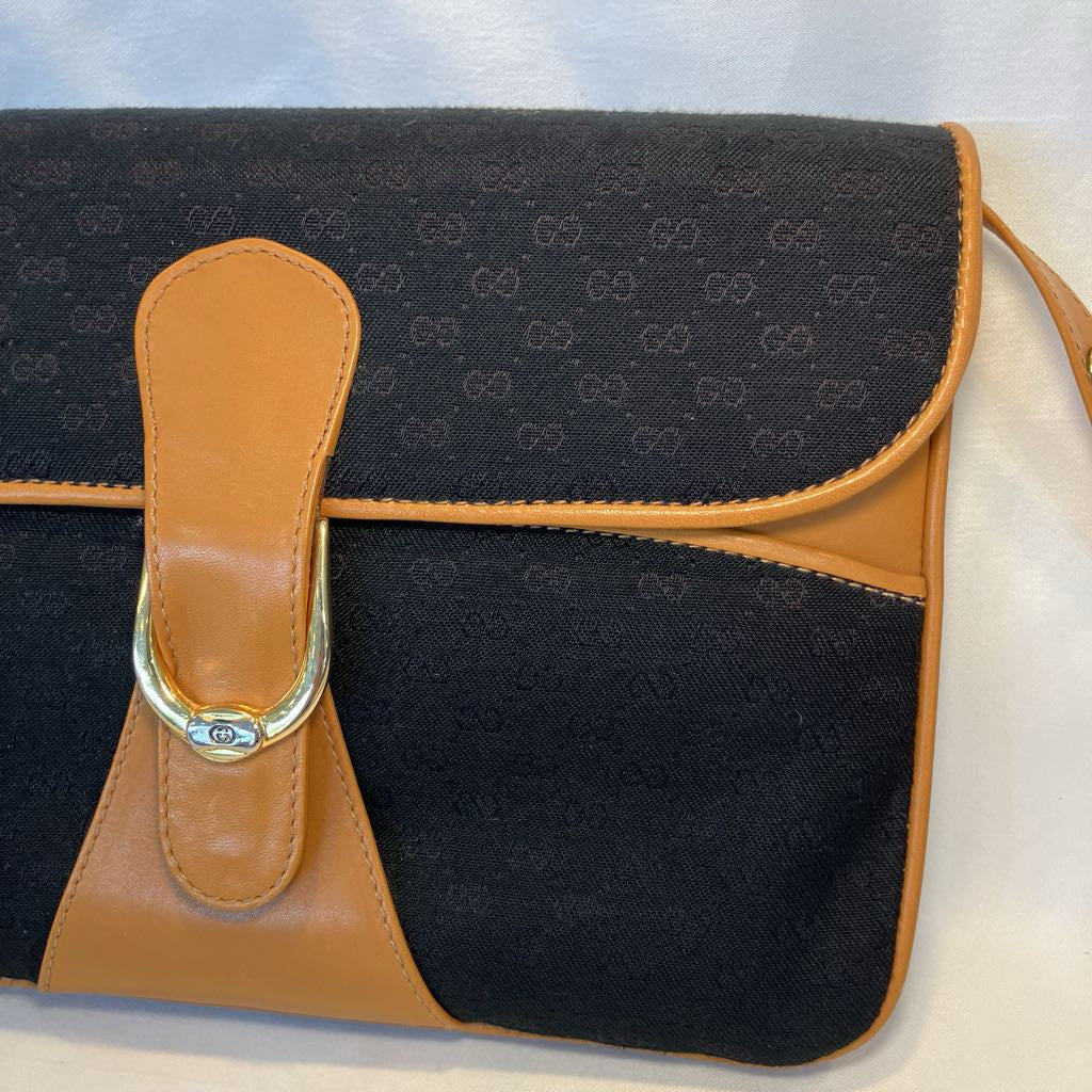 A close-up of a Gucci shoulder bag featuring the brand's signature GG monogram canvas in black and tan, with a tan leather strap closure and a gold-tone metal ring.