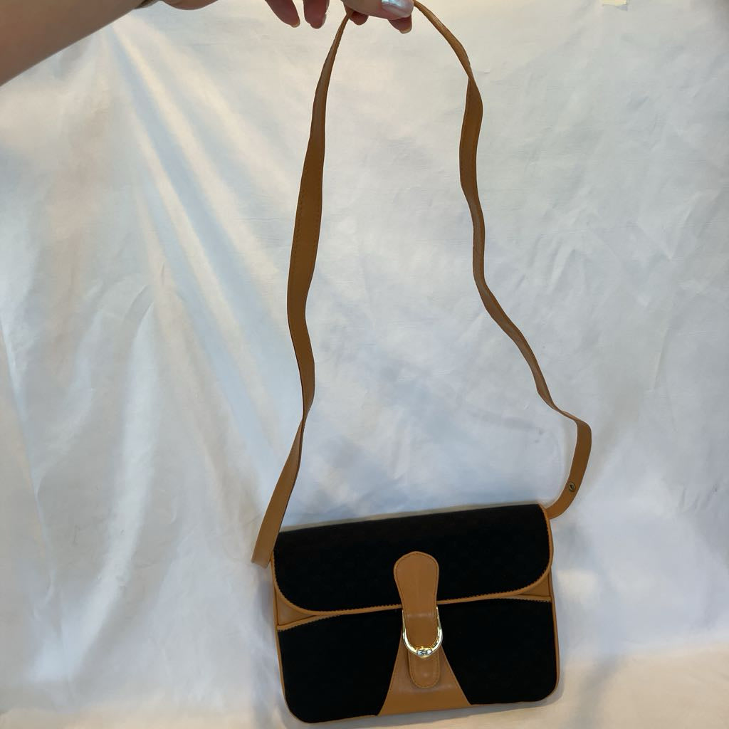 A black and tan shoulder bag with a long strap, held by a hand against a wrinkled white fabric background.