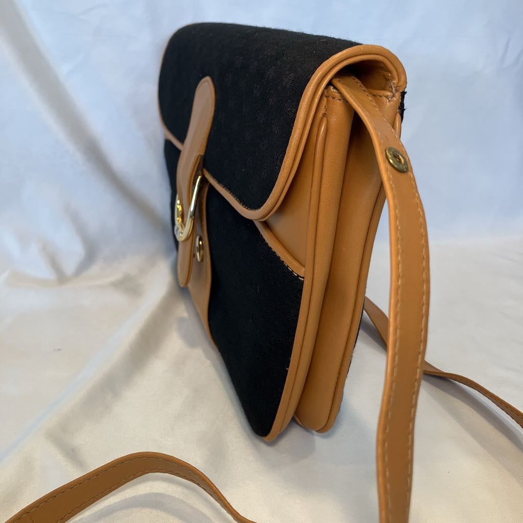 A black and tan crossbody bag with a black fabric body, tan leather trim, and a gold-tone buckle closure, shown on a white fabric background.