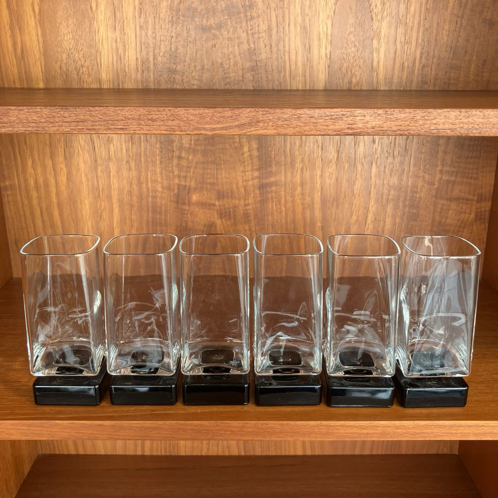 Set of six clear glass tumblers with black bases on a wooden shelf.