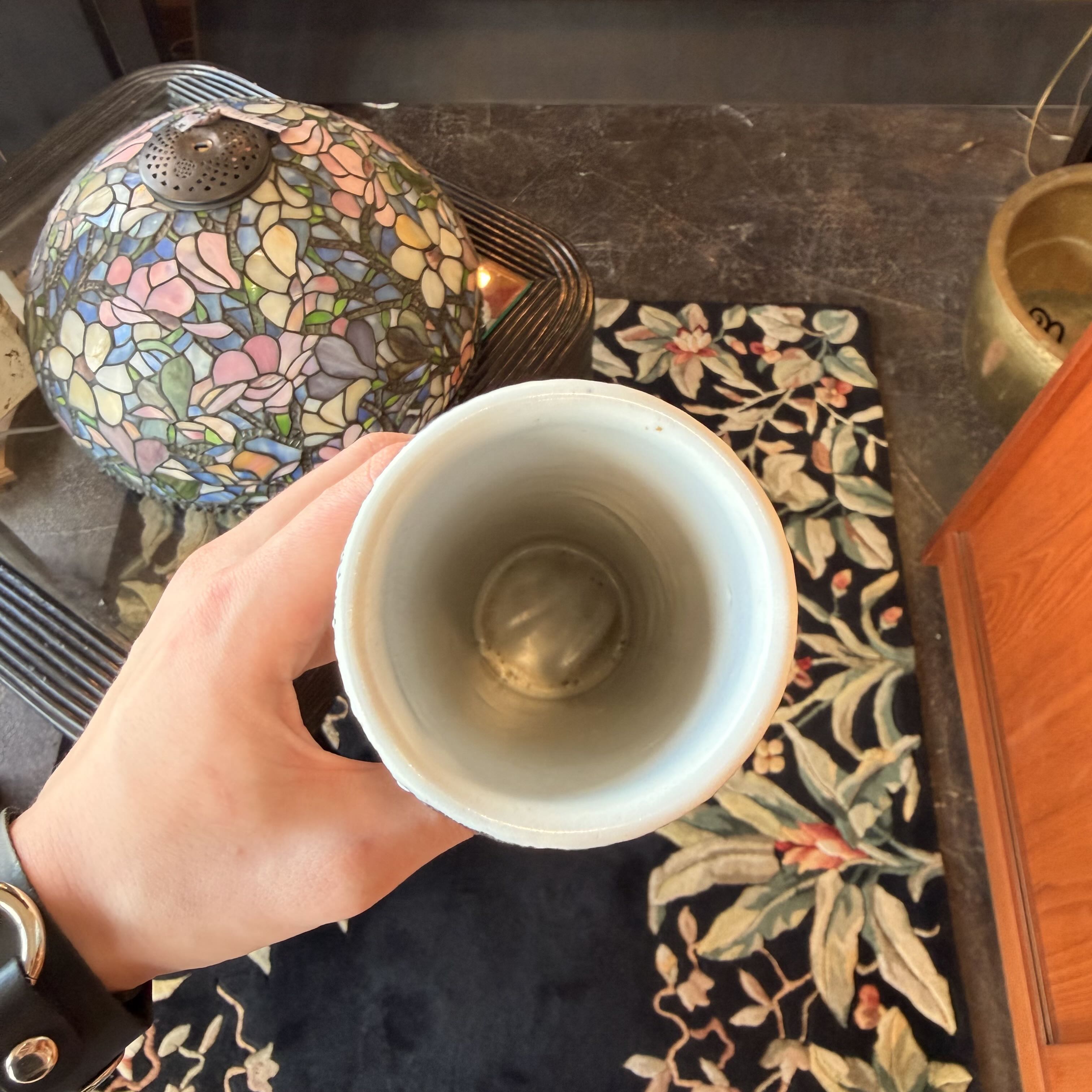 Hand holding a white ceramic cup with a decorative lamp and rug in the background