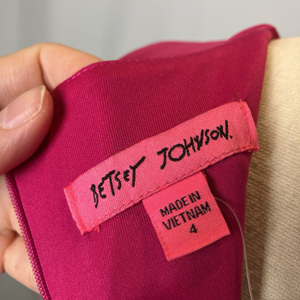 Close-up of a Betsey Johnson label on a pink garment with a beige background.