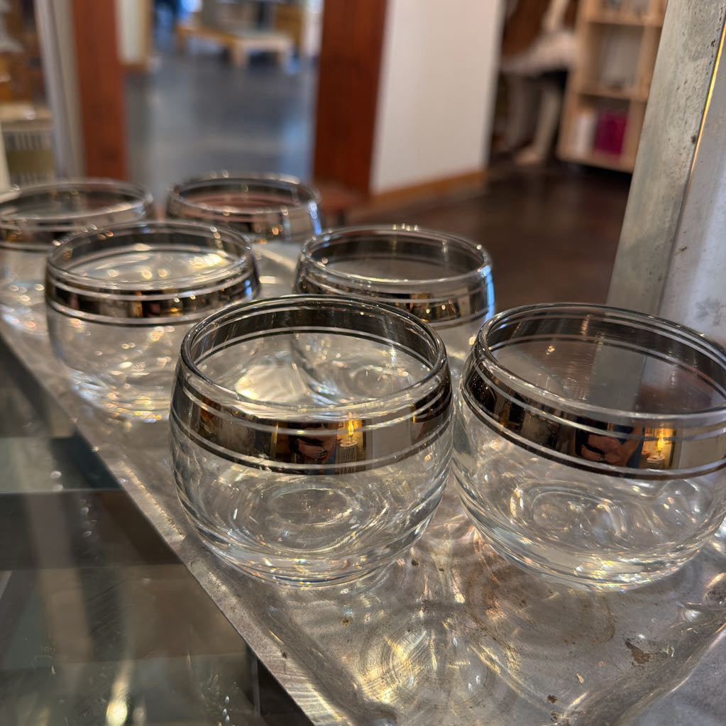 Five clear glass tumblers with metallic silver bands around the rims, arranged on a reflective metallic tray in an indoor setting.
