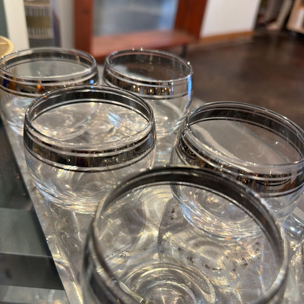 Five clear glass tumblers with metallic silver bands around the rims, arranged closely together on a reflective surface.