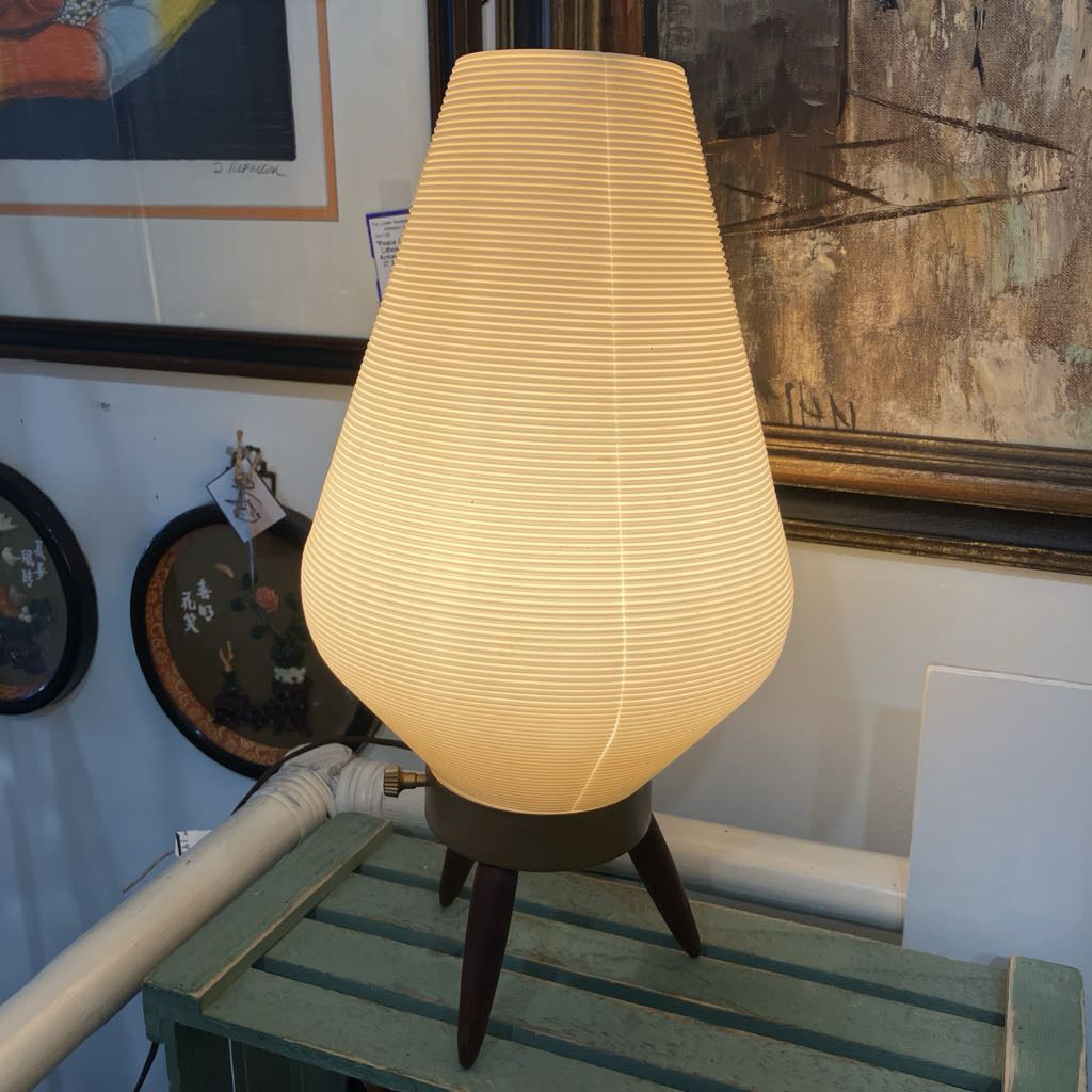 Lamp with a textured shade on a wooden surface, with artworks in the background