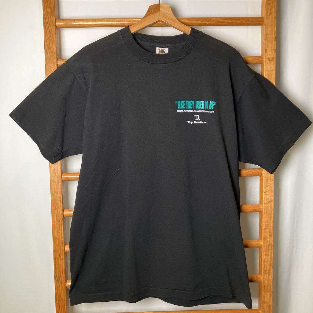 A black short-sleeve t-shirt hanging on a wooden hanger against a white curtain backdrop with a wooden ladder visible behind it. The shirt features a small graphic on the left chest with the text '"LIKE THEY USED TO BE"' in teal, followed by smaller white text, and a small logo below.