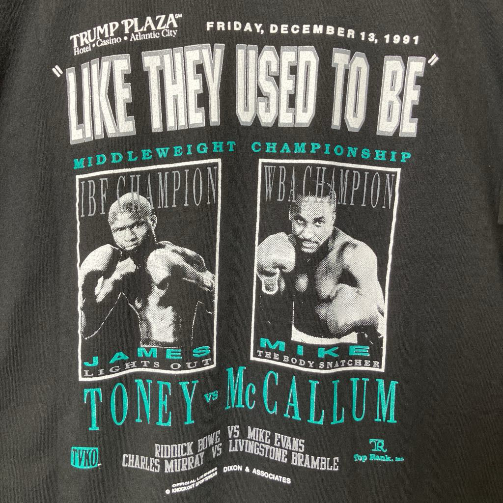 Close-up of a black graphic T-shirt featuring large white text reading "LIKE THEY USED TO BE" with two boxer portraits and turquoise text including "TONEY vs McCALLUM" and event details from Friday, December 13, 1991.
