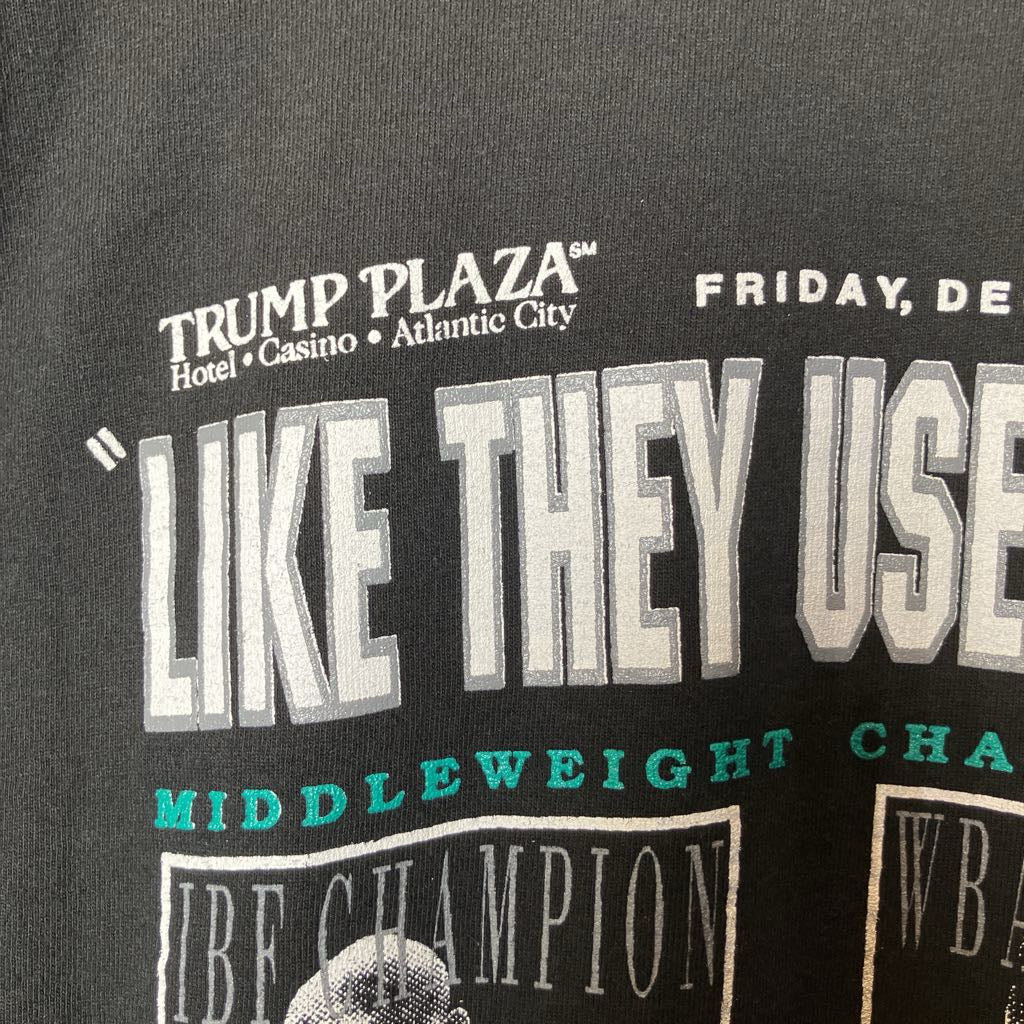 Close-up of a black graphic t-shirt featuring a distressed white and teal print with the text 'TRUMP PLAZA' and 'LIKE THEY USE'.