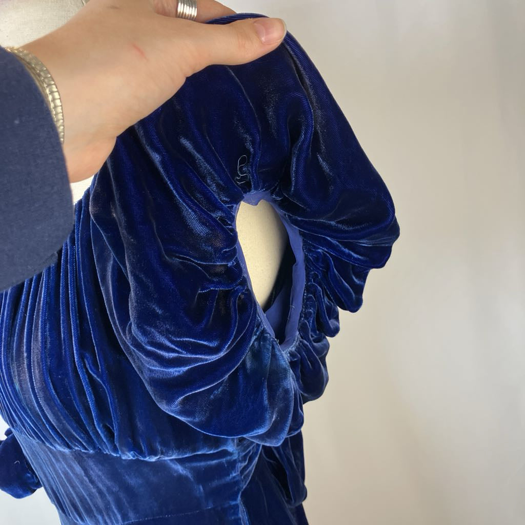 Blue velvet garment on a mannequin with a hand adjusting it against a neutral background