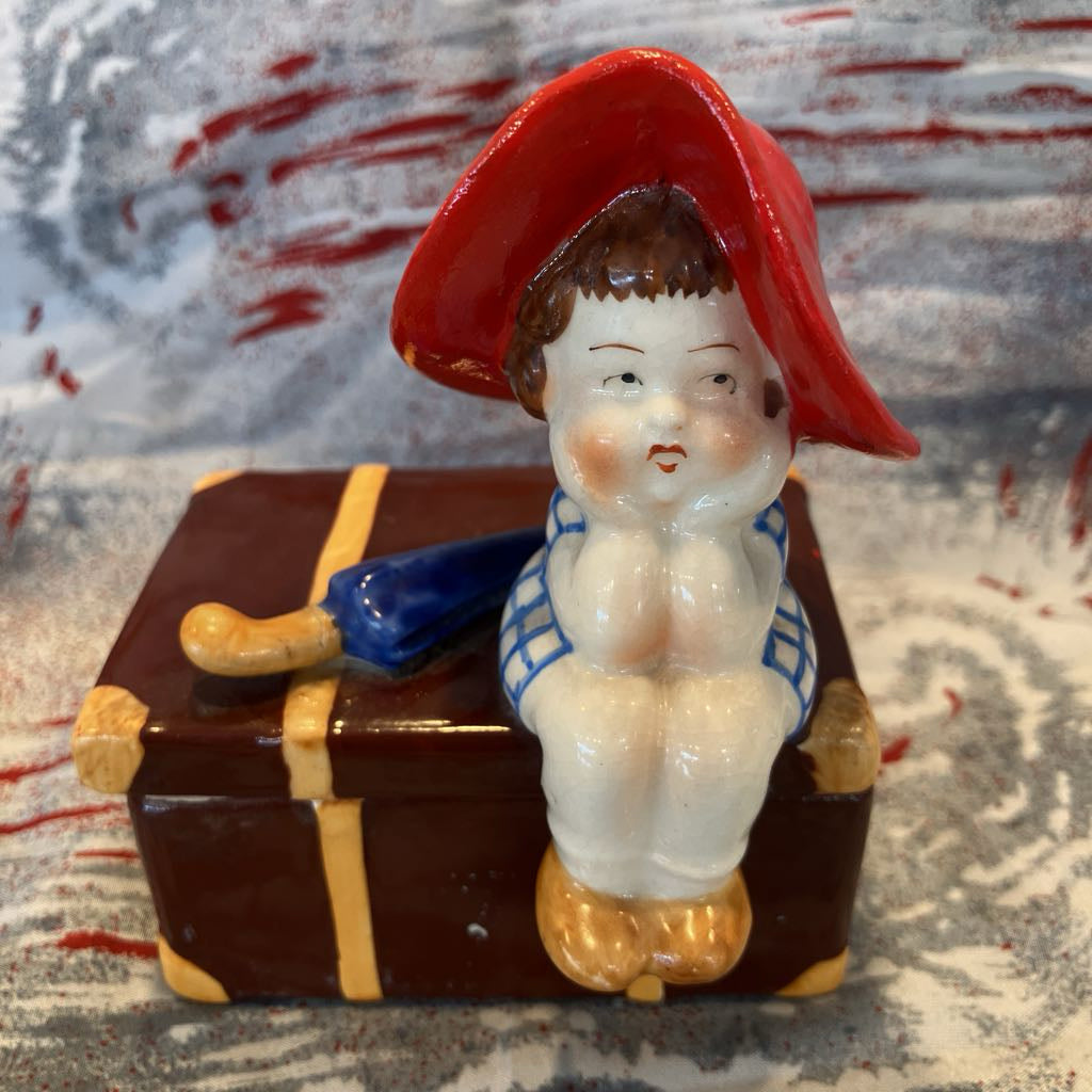 Small ceramic figurine of a child with a red hat sitting on a suitcase against a textured background