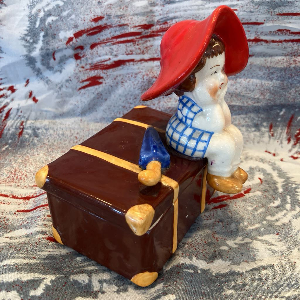 Small figurine of a child with a red hat sitting on a suitcase against a textured background