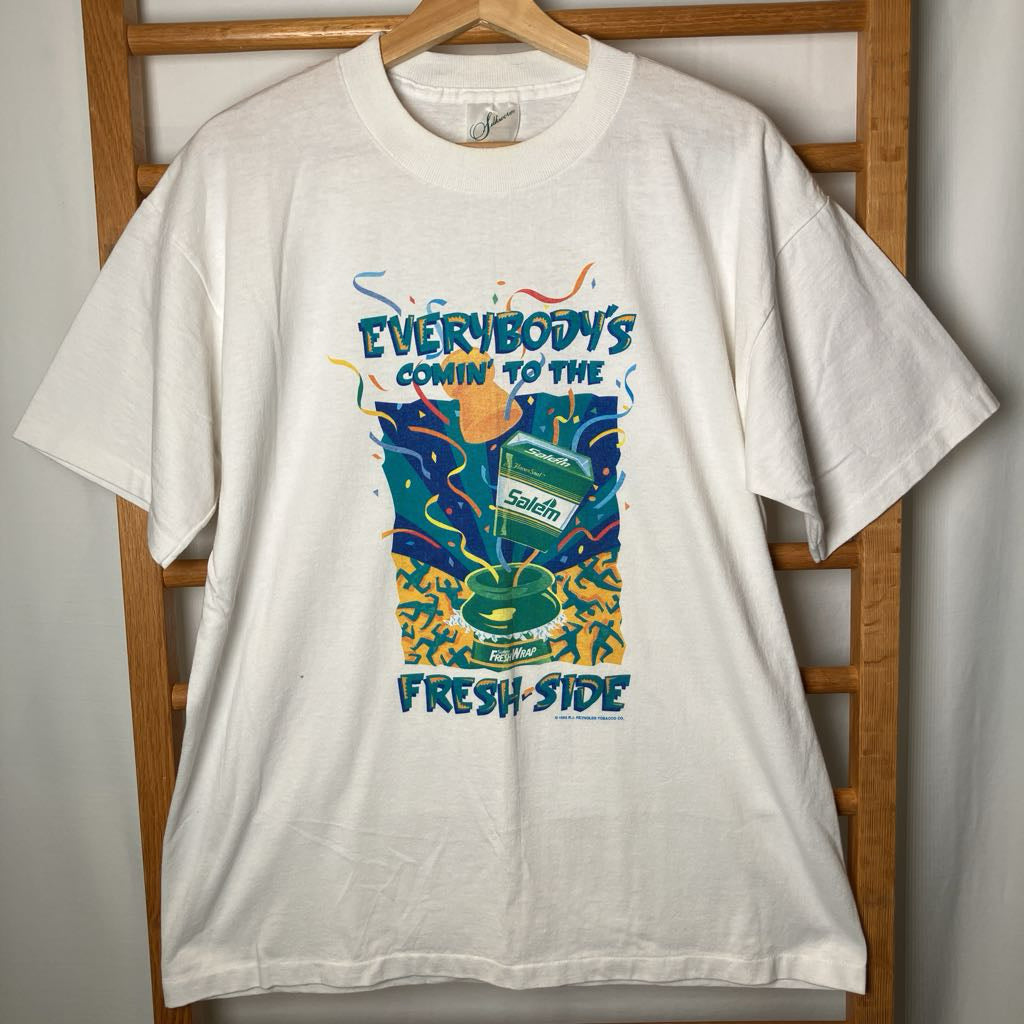 A white short-sleeve graphic t-shirt hanging on a wooden hanger against a wooden chair, featuring a colorful retro-style print with the text 'EVERYBODY'S COMIN' TO THE Fresh Side' and a cassette tape illustration.