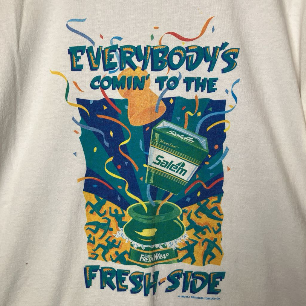 A close-up of a cream-colored t-shirt featuring a colorful vintage-style graphic print. The graphic displays a green carton labeled 'Saflem' pouring into a green cup, surrounded by colorful confetti and the text 'EVERYBODY'S COMIN' TO THE FRESH SIDE'.