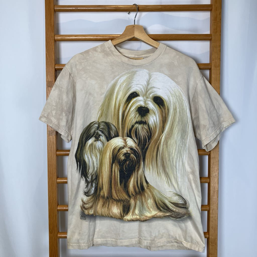 Beige t-shirt with a graphic of three dogs on a wooden hanger against a white wall.