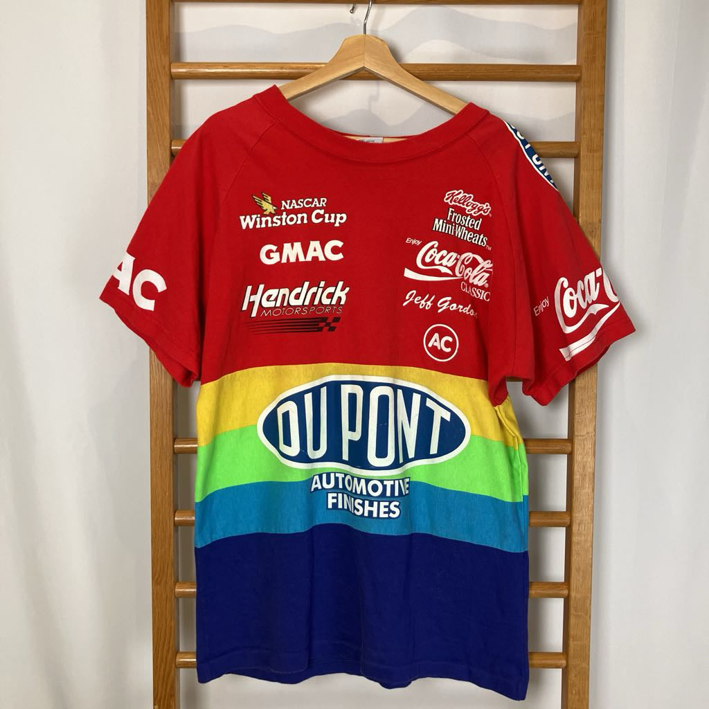 Red t-shirt with multiple brand logos on a wooden hanger against a white wall.