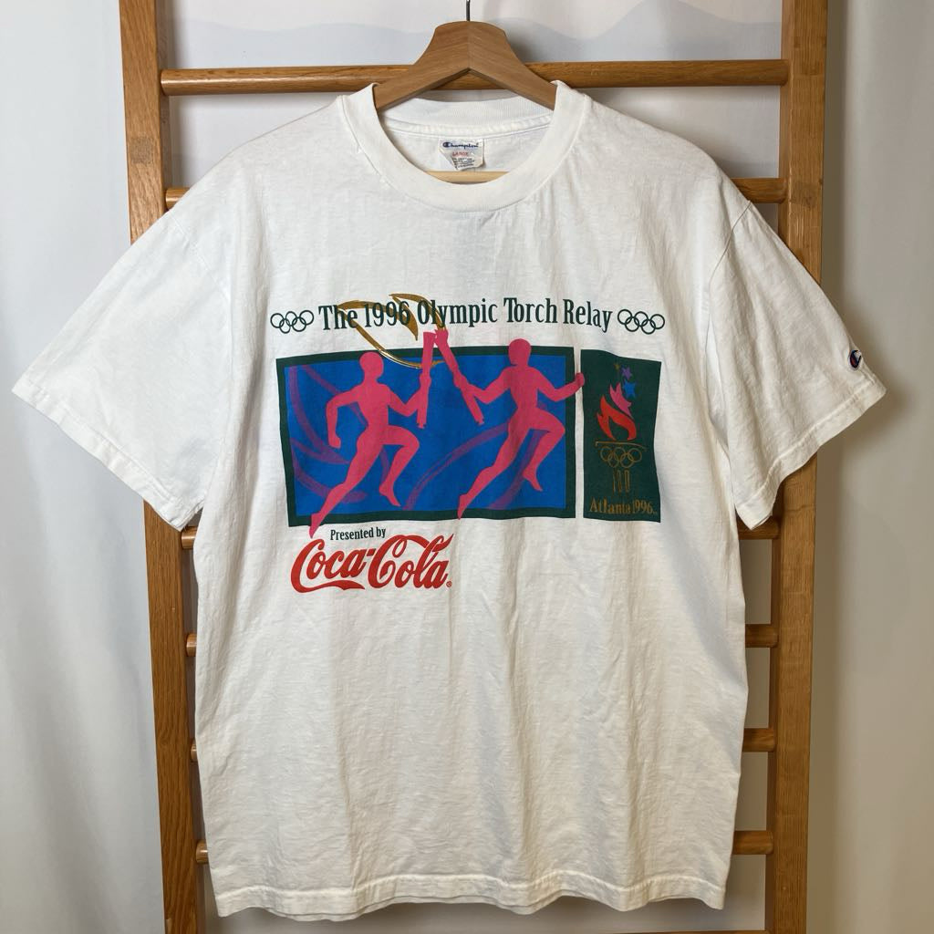 White t-shirt with Olympic torch relay graphic and Coca-Cola logo on a wooden hanger.