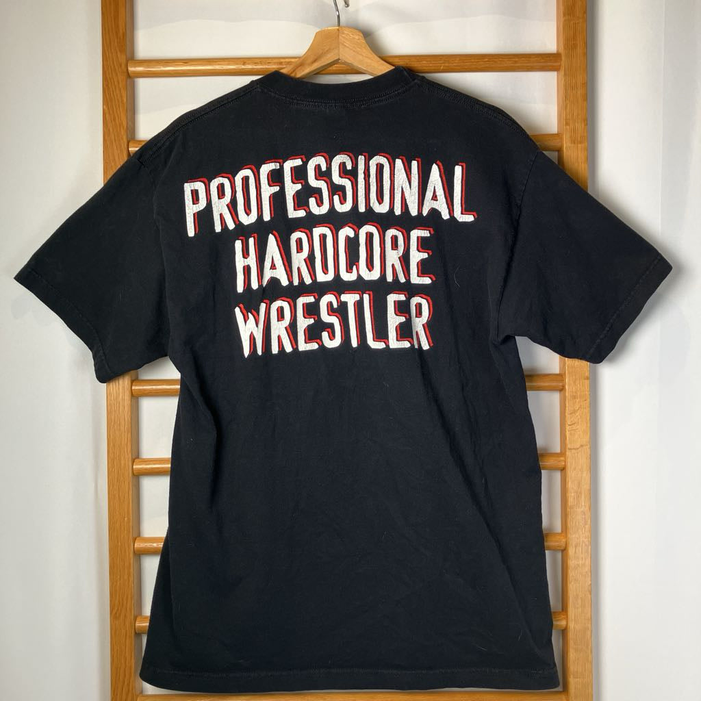 Black t-shirt with 'Professional Hardcore Wrestler' text on a wooden hanger against a white wall.