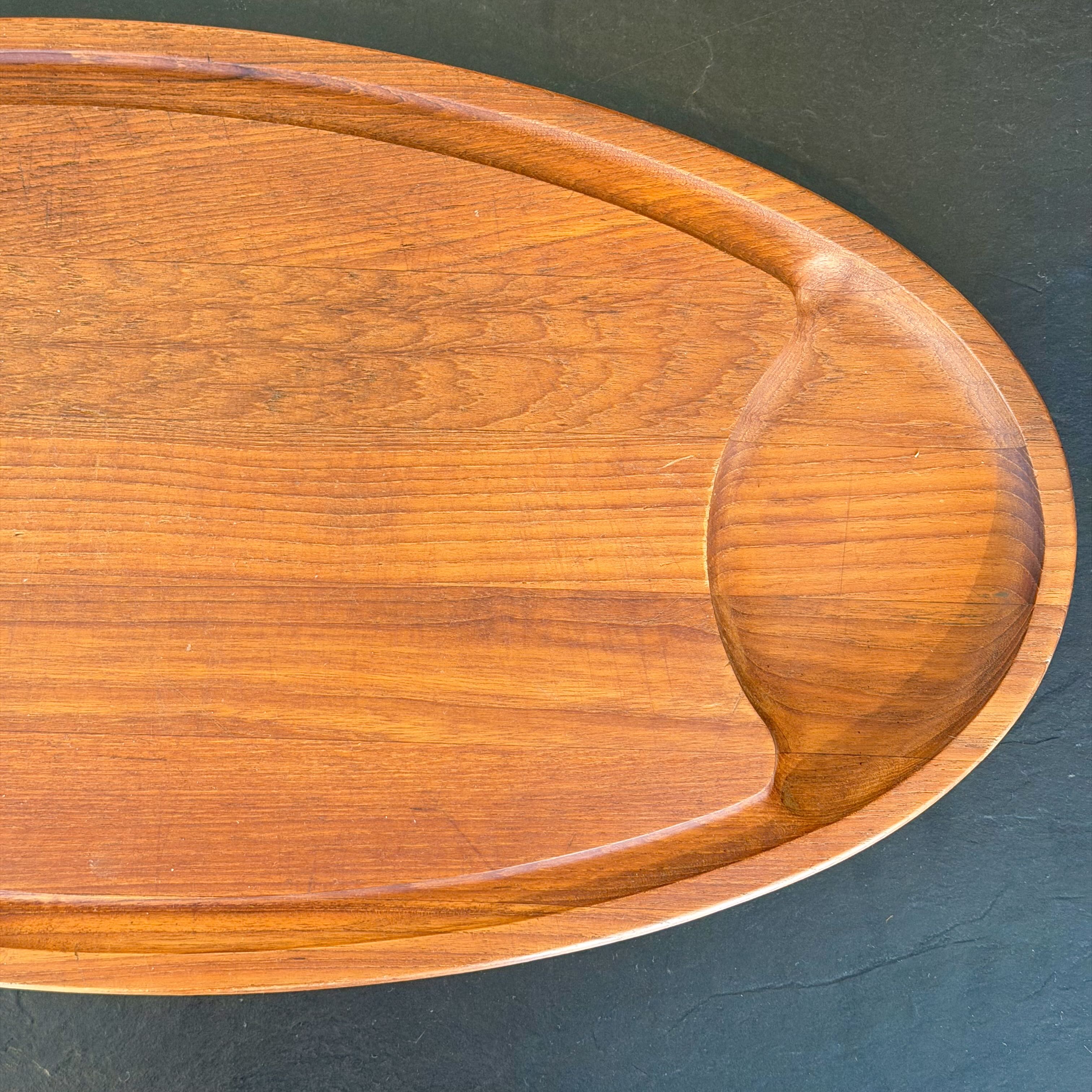 An oval wooden tray with a warm honey-brown finish and visible wood grain, photographed close-up against a dark textured surface.