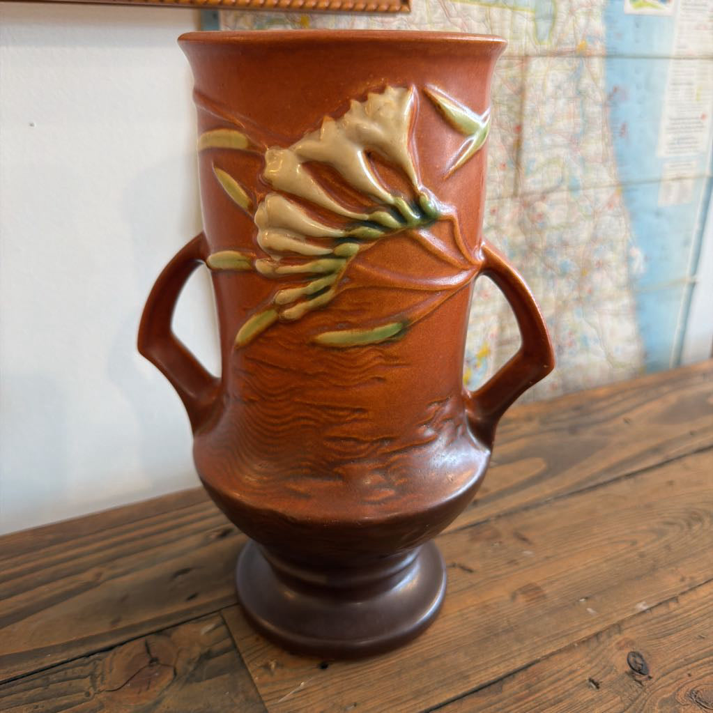 A vintage terracotta-colored ceramic vase with two side handles, featuring raised floral relief decoration in cream and green, displayed on a wooden surface.