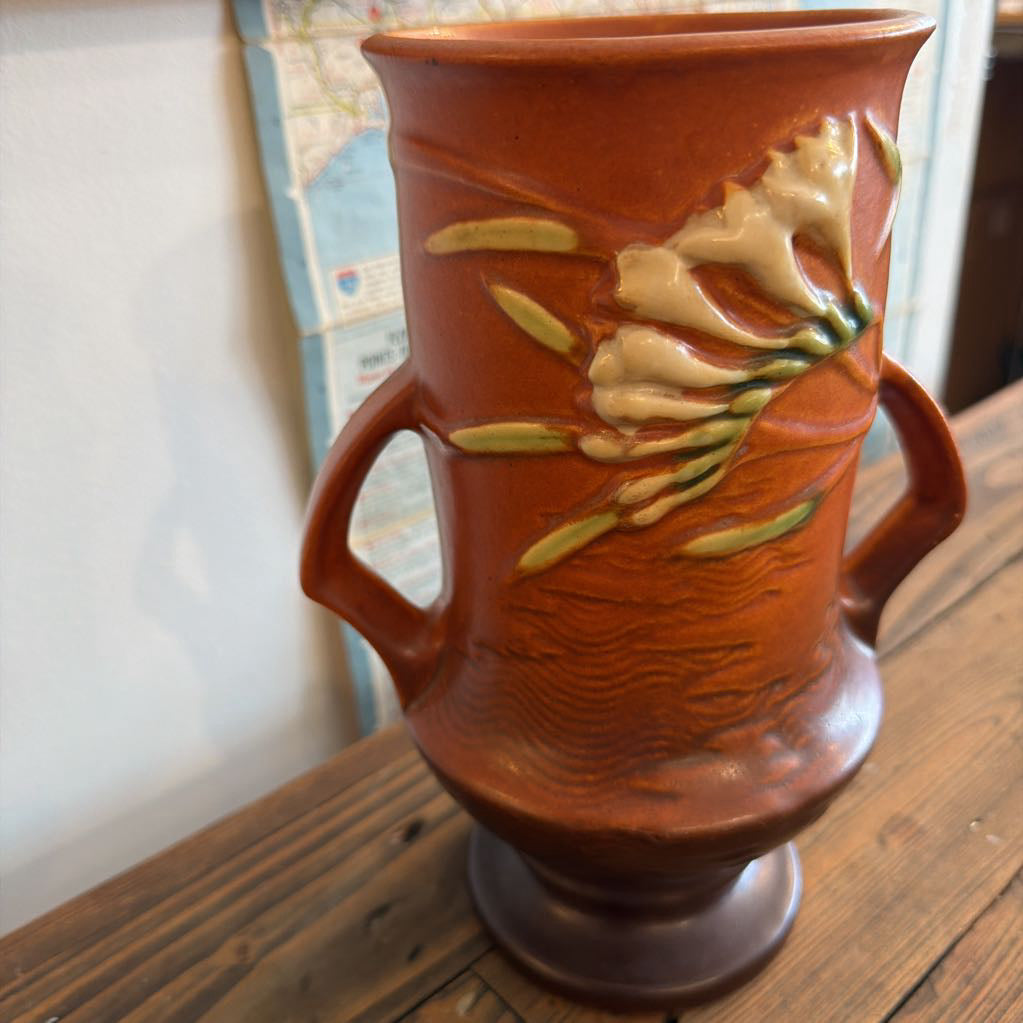 A terracotta-colored ceramic vase with two side handles, featuring raised cream-colored floral motifs and green leaves on the front, displayed on a wooden surface.