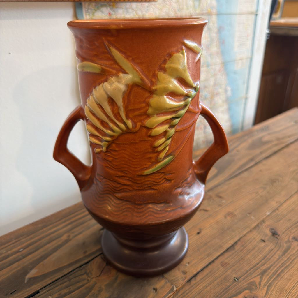 A terracotta-colored ceramic vase with two side handles and raised seashell and wave-like relief details, displayed on a wooden table.