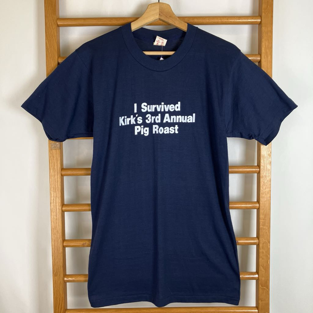 A navy blue short-sleeve t-shirt hanging on a wooden hanger against a light-colored wall, with white text on the chest that reads 'I Survived Kirk's 3rd Annual Pig Roast'.