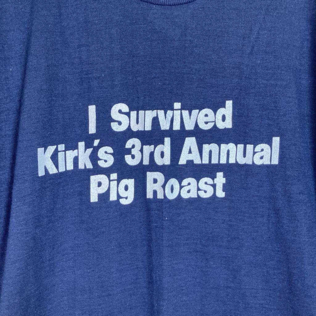 A close-up of a heather blue t-shirt featuring the text 'I Survived Kirk's 3rd Annual Pig Roast' printed in white across the chest.