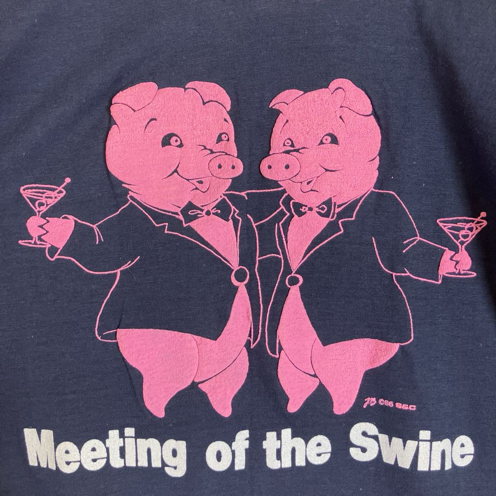 Close-up of a navy blue graphic t-shirt featuring two pink cartoon pigs in black tuxedos holding martini glasses, with the text 'Meeting of the Swine' printed in white below.
