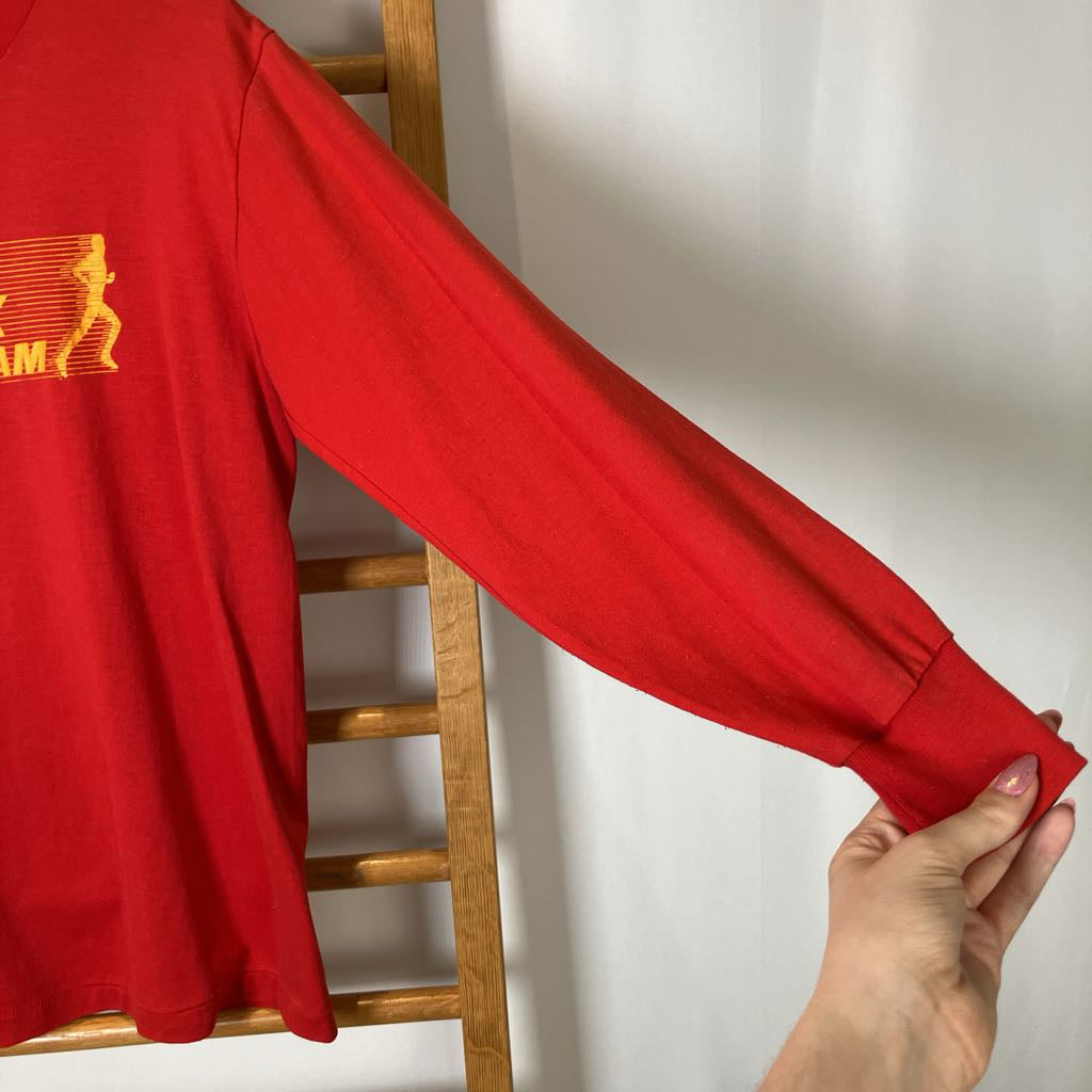 Red sweatshirt with a yellow logo held by a hand against a white wall.