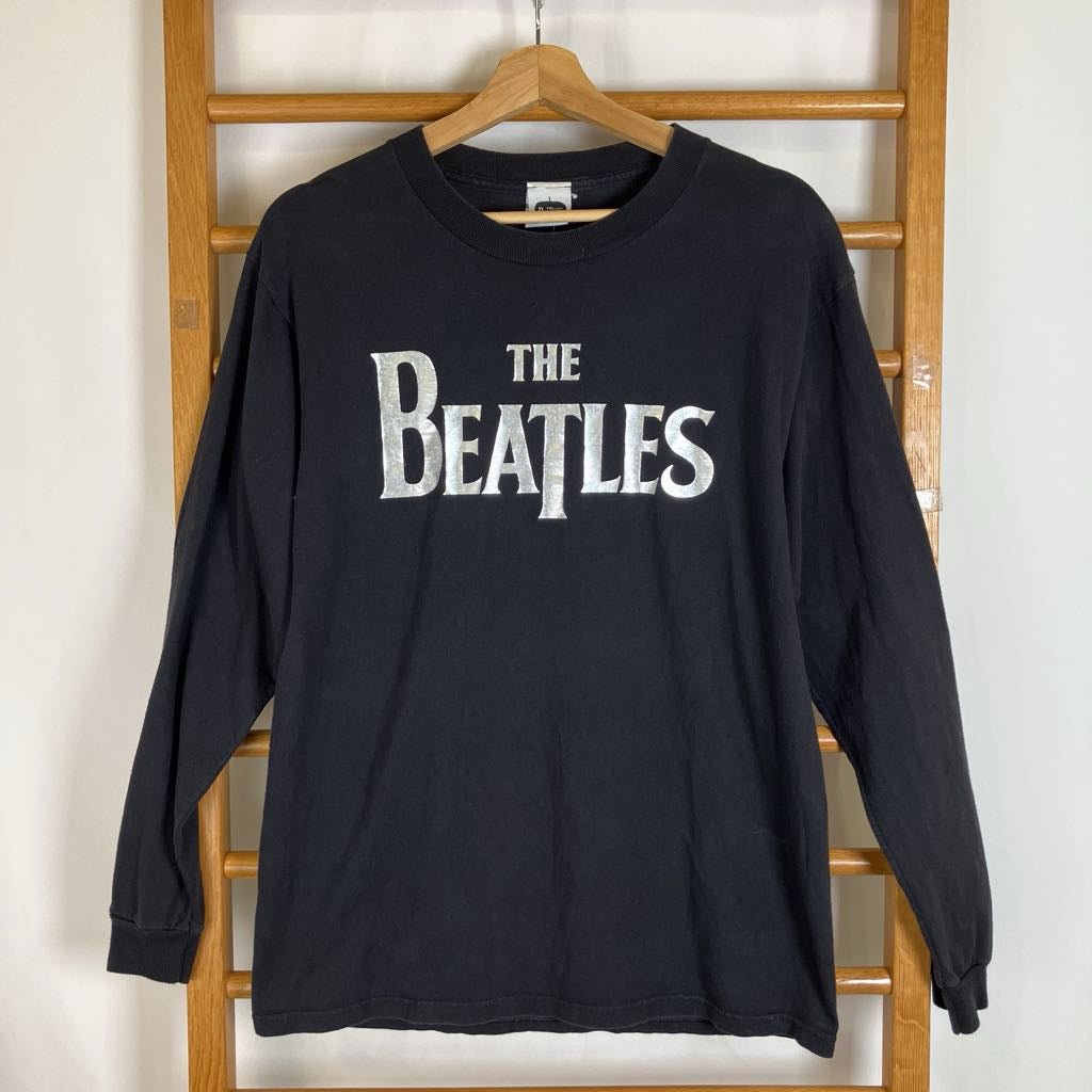 A black long-sleeve crewneck t-shirt featuring the white distressed 'THE BEATLES' logo across the chest, hanging on a wooden hanger from a wooden clothes rack against a white wall.