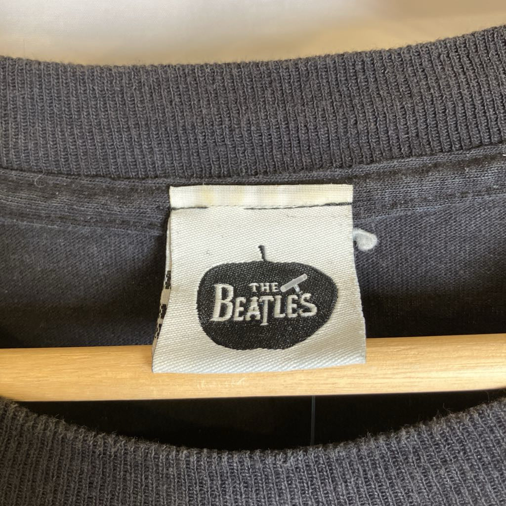 Close-up of a dark gray ribbed knit garment on a wooden hanger, showing a white fabric tag with a black apple-shaped logo that reads 'The Beatles'.