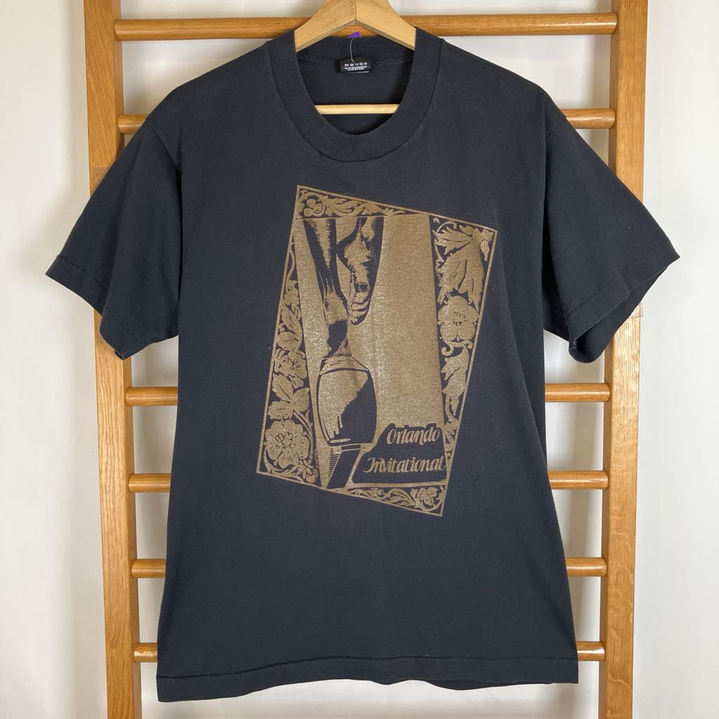 A dark charcoal gray short-sleeve t-shirt with a vintage-style graphic print on the front, hanging on a wooden hanger from a wooden ladder against a white wall.