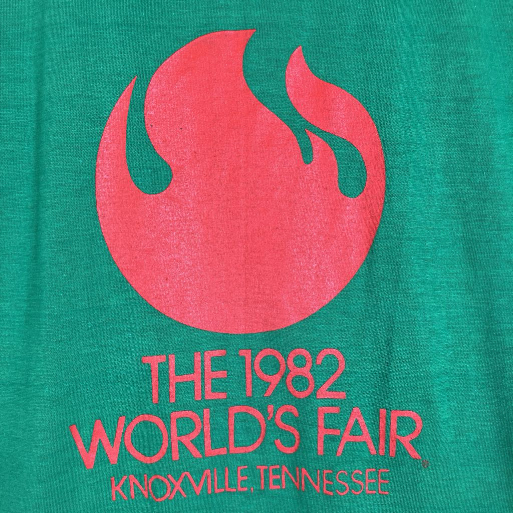A close-up of a green t-shirt featuring a large red graphic of a flame-like shape above the text 'THE 1982 WORLD'S FAIR KNOXVILLE TENNESSEE' in red.
